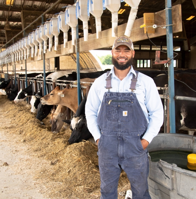 A farmer standing in a barn. Behind him are cows in stalls, eating hay.